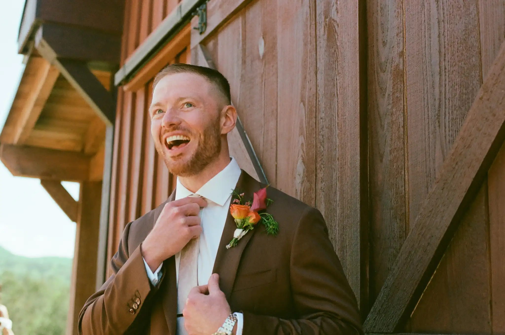 Groom in a brown suit laughing as he straightens his tie by the barn door, shot on 35mm film at The Venue at Sharp Mountain in Jasper, Georgia