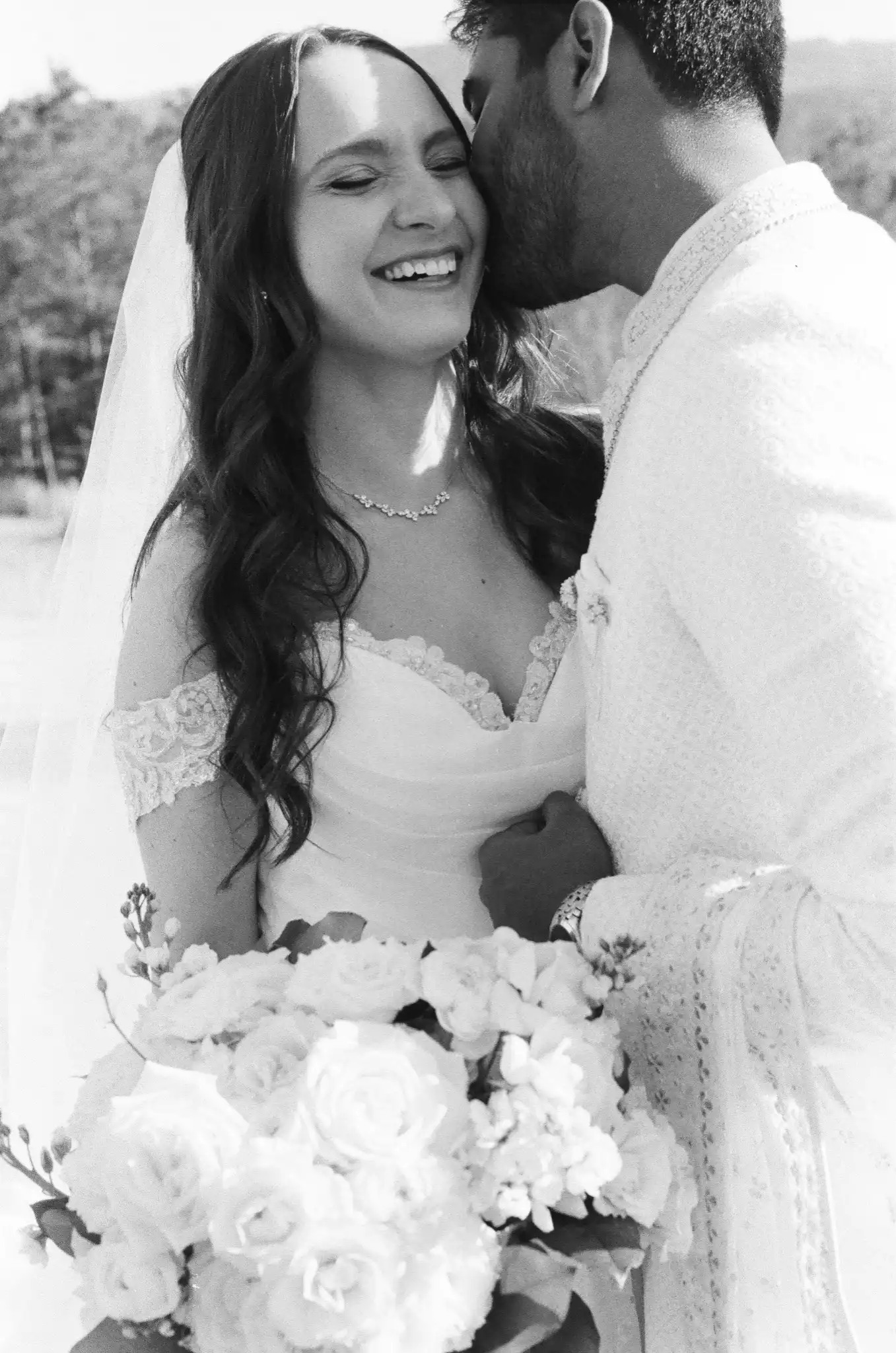 Groom in a sherwani kissing the laughing bride's cheek as she holds white roses, in black and white on 35mm film at The Venue at Sharp Mountain in Jasper, Georgia