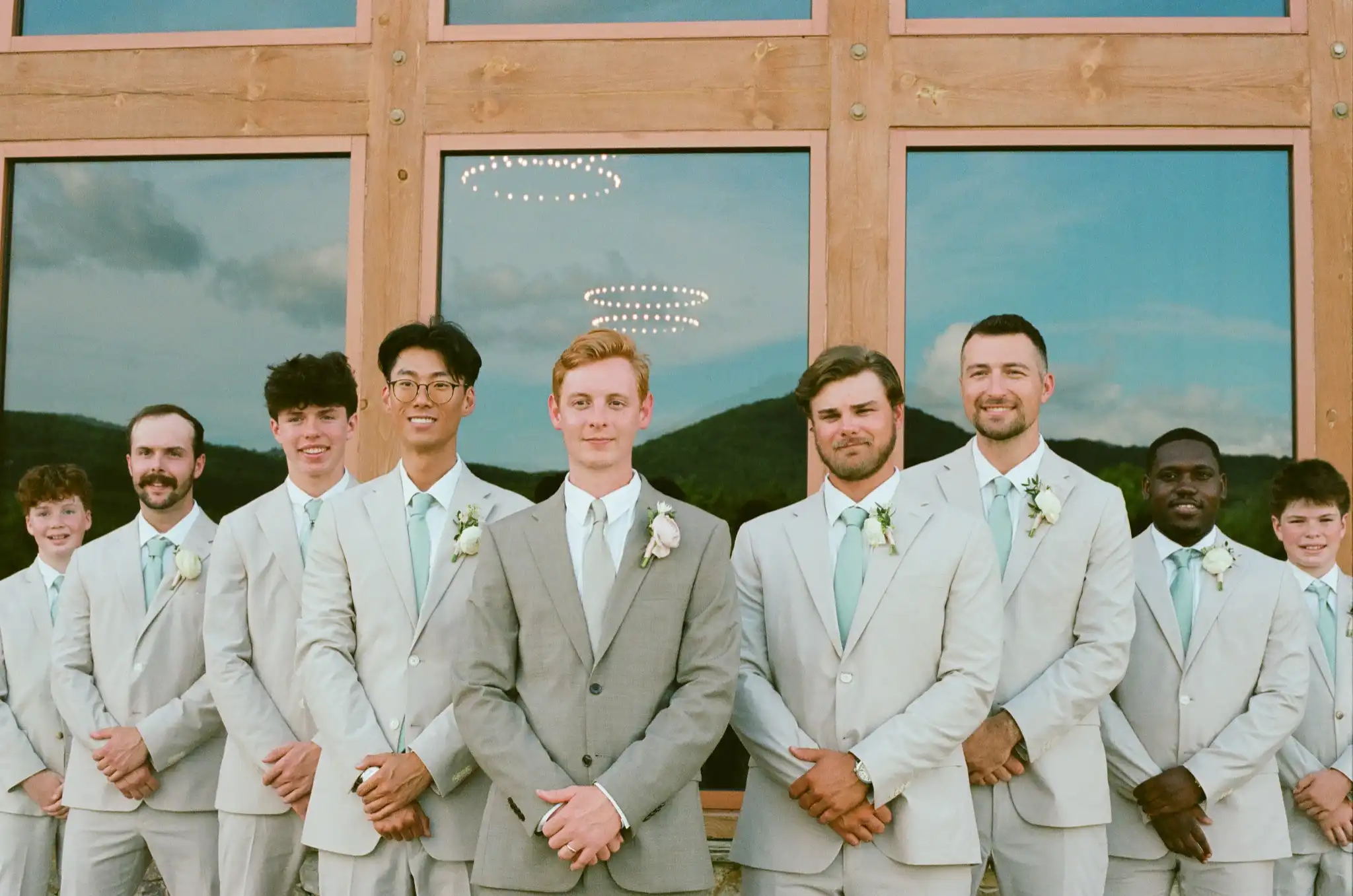 Groom and his groomsmen in tan suits with sage ties by the barn windows, shot on 35mm film at The Venue at Sharp Mountain in Jasper, Georgia