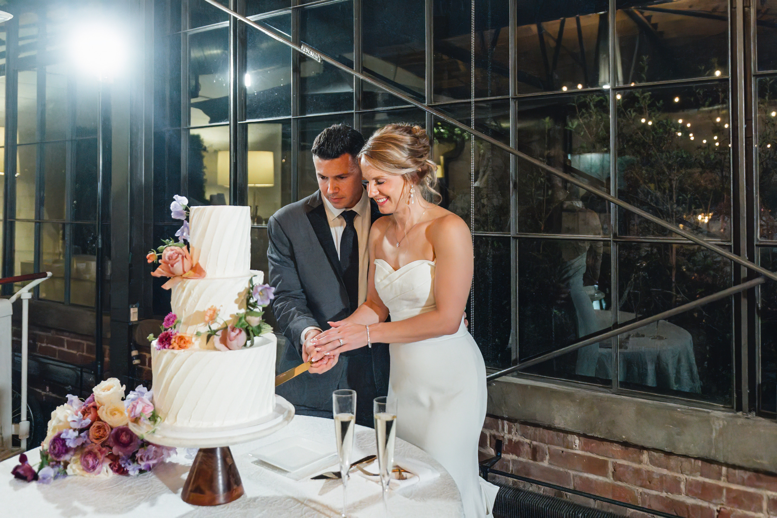 Wedding Reception: cutting the cake against the black steel windows, at their wedding at Summerour Studio in Atlanta, GA