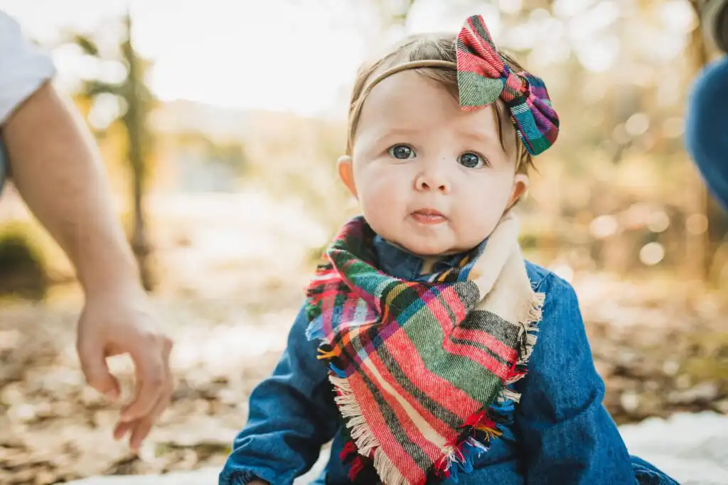Baby in a bow and plaid scarf photographed outdoors in autumn leaves during a portrait session